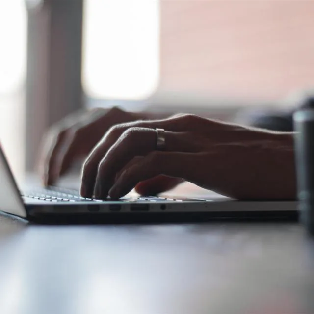 Hands at a laptop keyboard mid-sentence, a notebook open alongside.