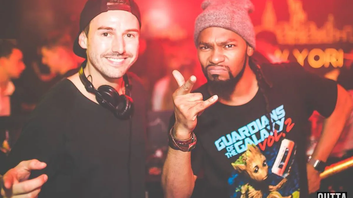 Two performers lit red in a crowded club, one wearing headphones around his neck.