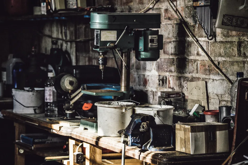 A working bench crowded with tools, a drill press and jars of parts, lit
                  from one side.