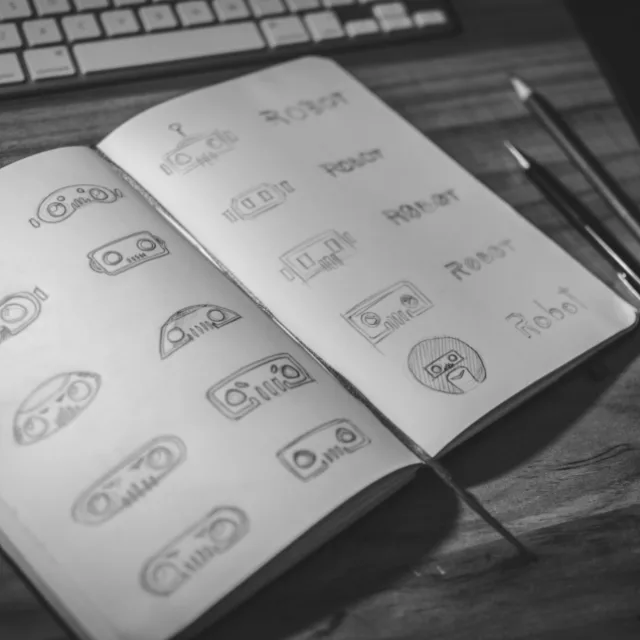 An open sketchbook filled with hand-drawn logo variations, a pencil and a keyboard beside it.