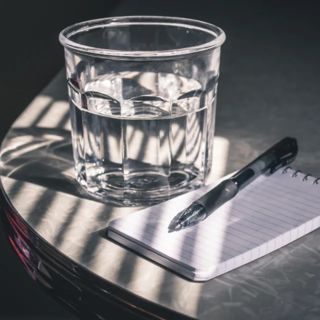 A small notepad and pen on a table beside a glass of water, in raking light.