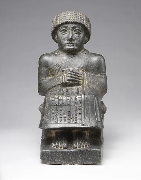 A seated diorite statue of a man with clasped hands and a brimmed woollen
                  cap, rows of cuneiform carved across his lap. Gudea of Lagash, named in
                  his own inscription as the man who built the temple.