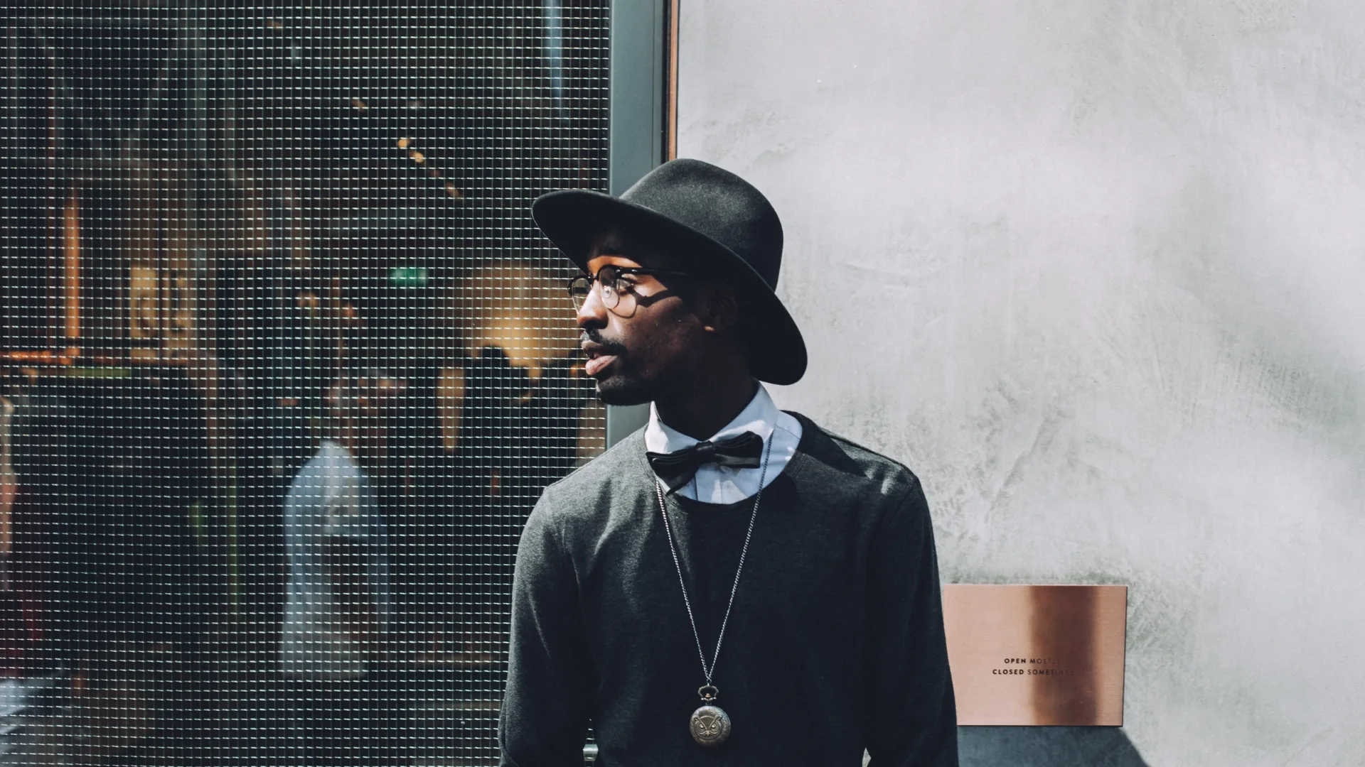A formally dressed man in a black hat standing beside a reflective glass wall.