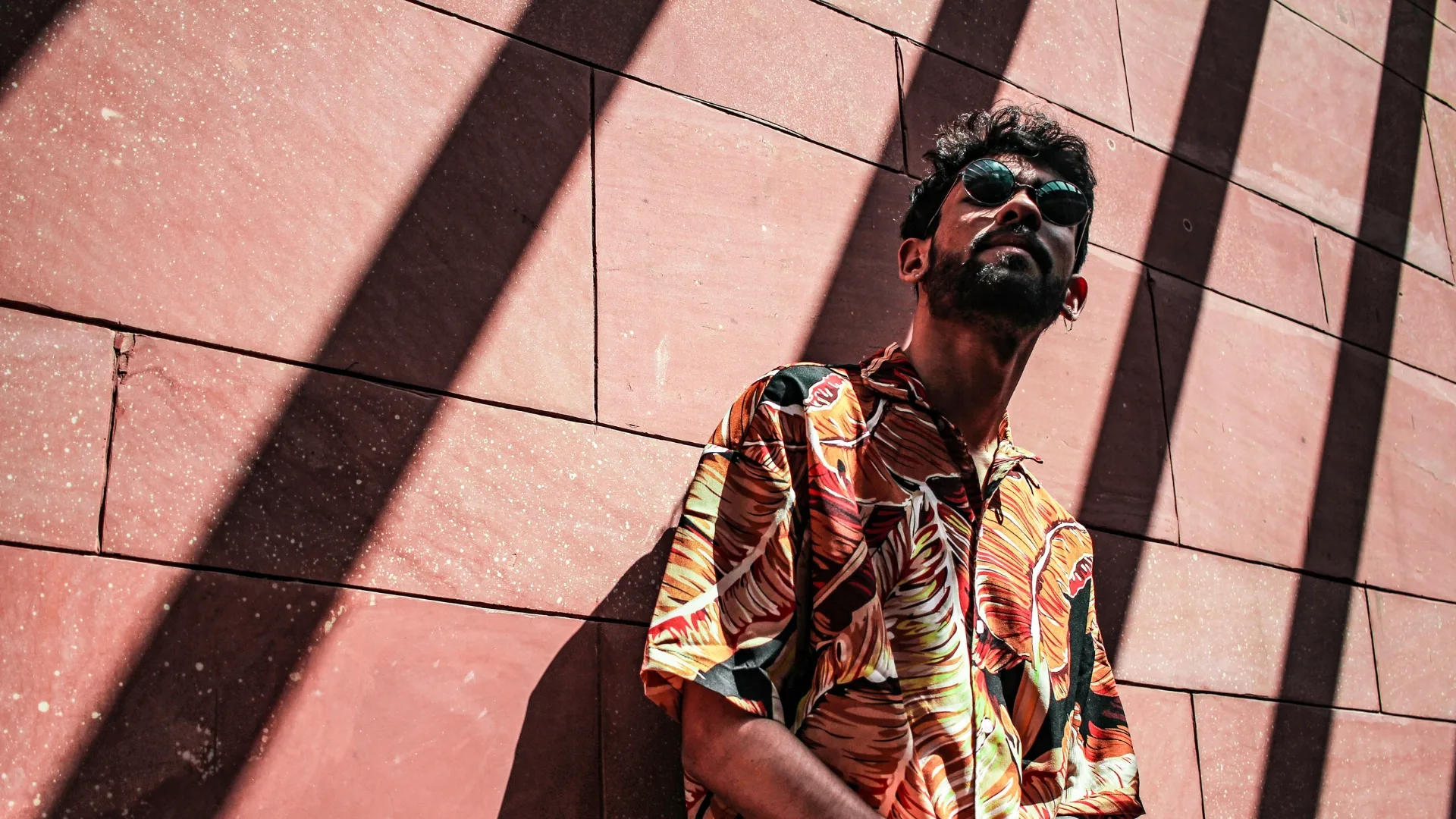 A man in sunglasses and a patterned shirt standing against a sunlit wall.