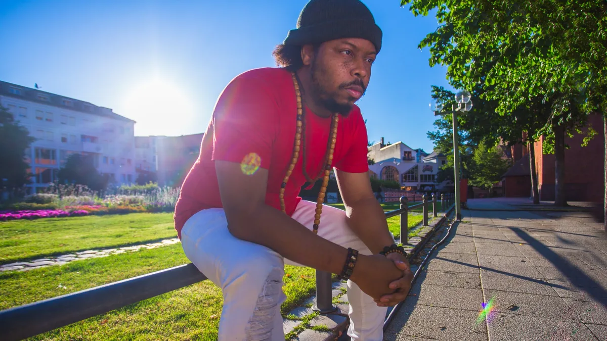 A man in a red shirt and headwrap crouching on a sunlit path, the low sun flaring behind him.