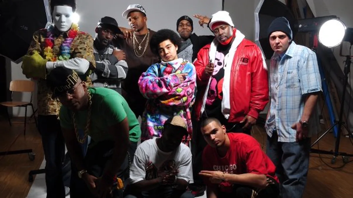 A group portrait of eleven performers under studio lights, standing and crouching in two rows.