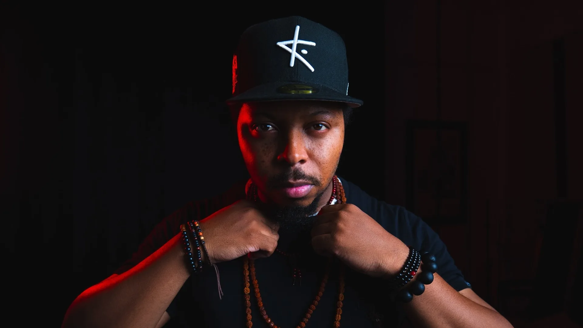 A studio portrait of a man in a black cap adjusting his collar under red rim light.